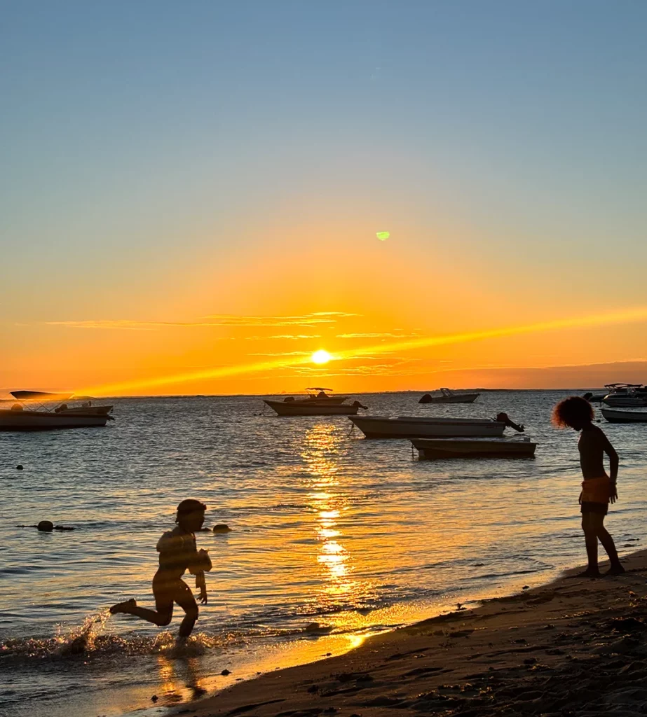 Deux enfants jouent dans l’eau à la plage publique de La Preneuse, avec pirogues sur le lagon et coucher de soleil à l’île Maurice